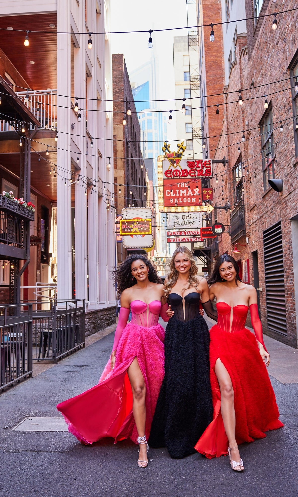 Three women in elegant prom ball gowns, including the Bustier Colette Long A-Line Prom Ball Gown CL5114, walk arm-in-arm down an urban alley lit with string lights and neon signs, smiling at the camera.